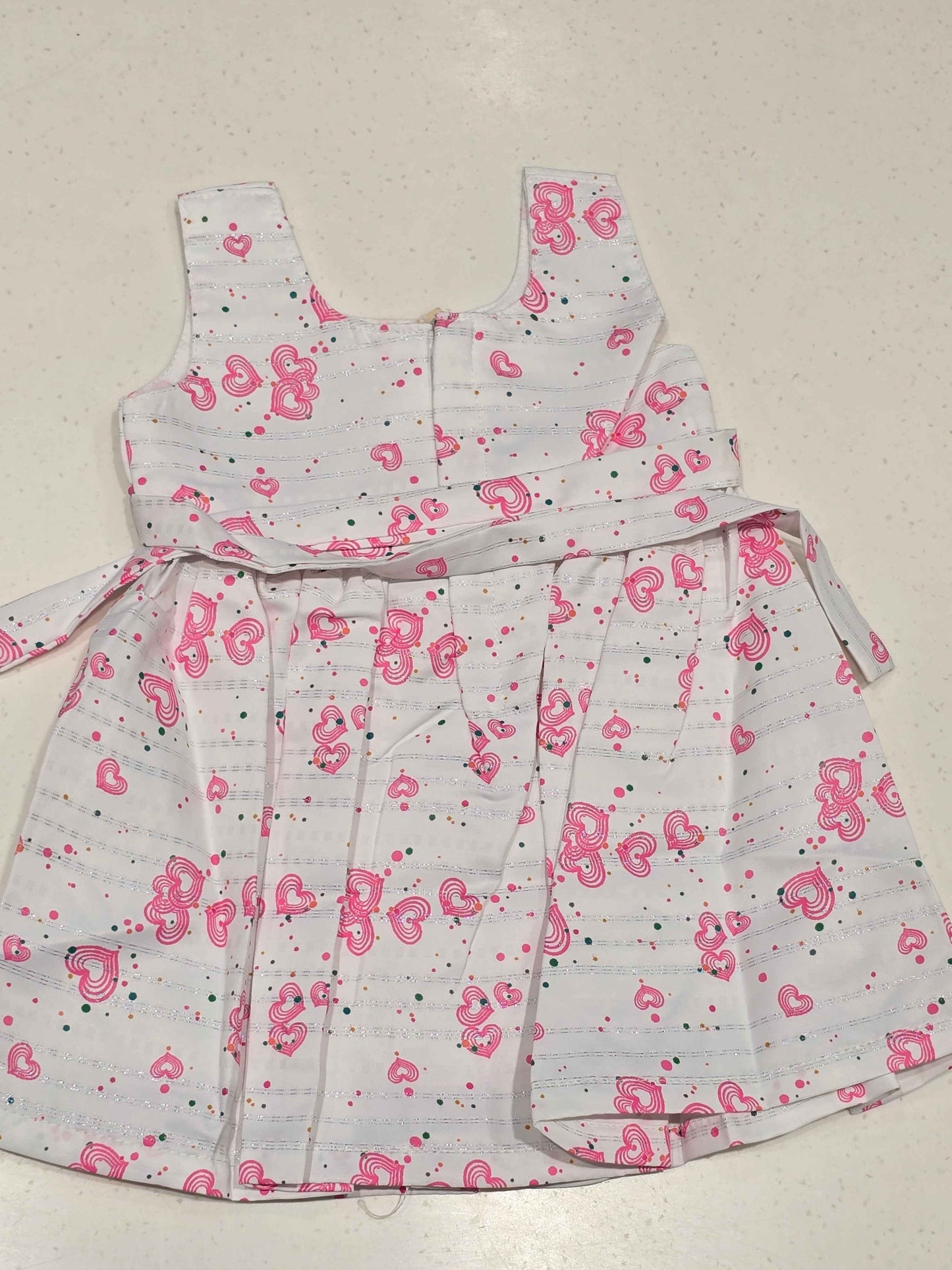 sleeveless, white baby girl cotton frock accented by a vibrant pink heart-shaped pattern and a centered waist bow