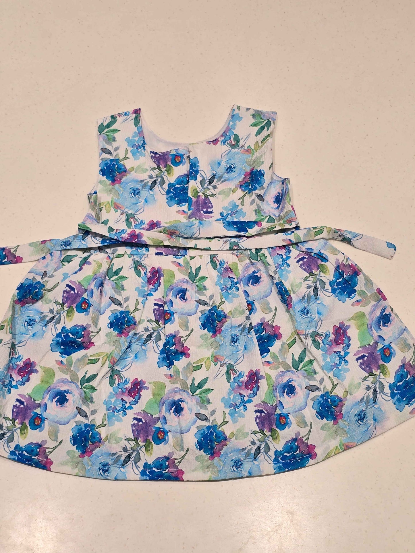 baby girl sleeveless cotton frock featuring a vibrant blue, purple, and green watercolor floral print on a crisp white background.