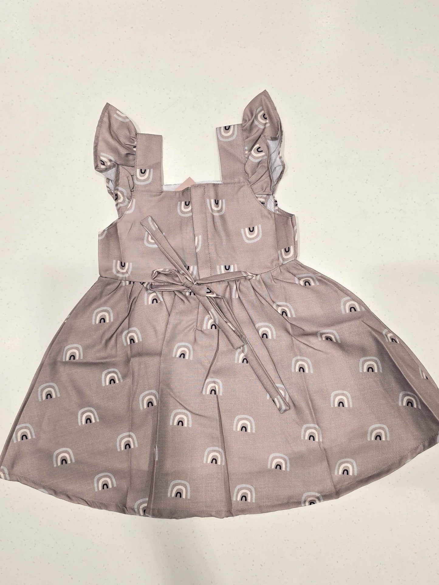 baby girl’s sleeveless cotton frock featuring a flared skirt, a gathered waist, square neckline, shoulder flutter ruffles, and a button-down back closure. It