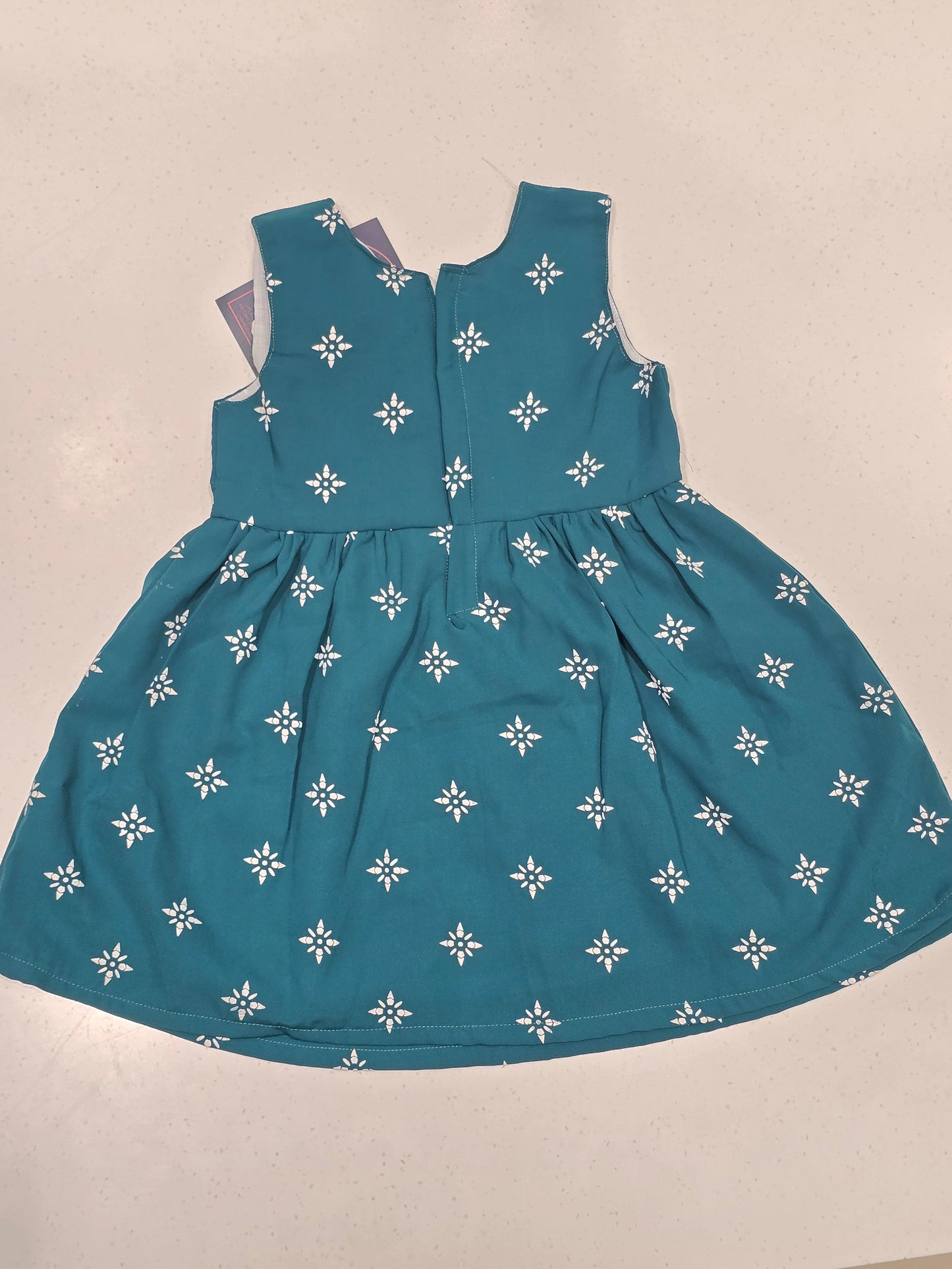 This teal cotton frock dress for girls features a classic dual-pattern design, with small white stars scattered across the sleeveless bodice and a geometric snowflake or floral print adorning the gathered A-line skirt. It