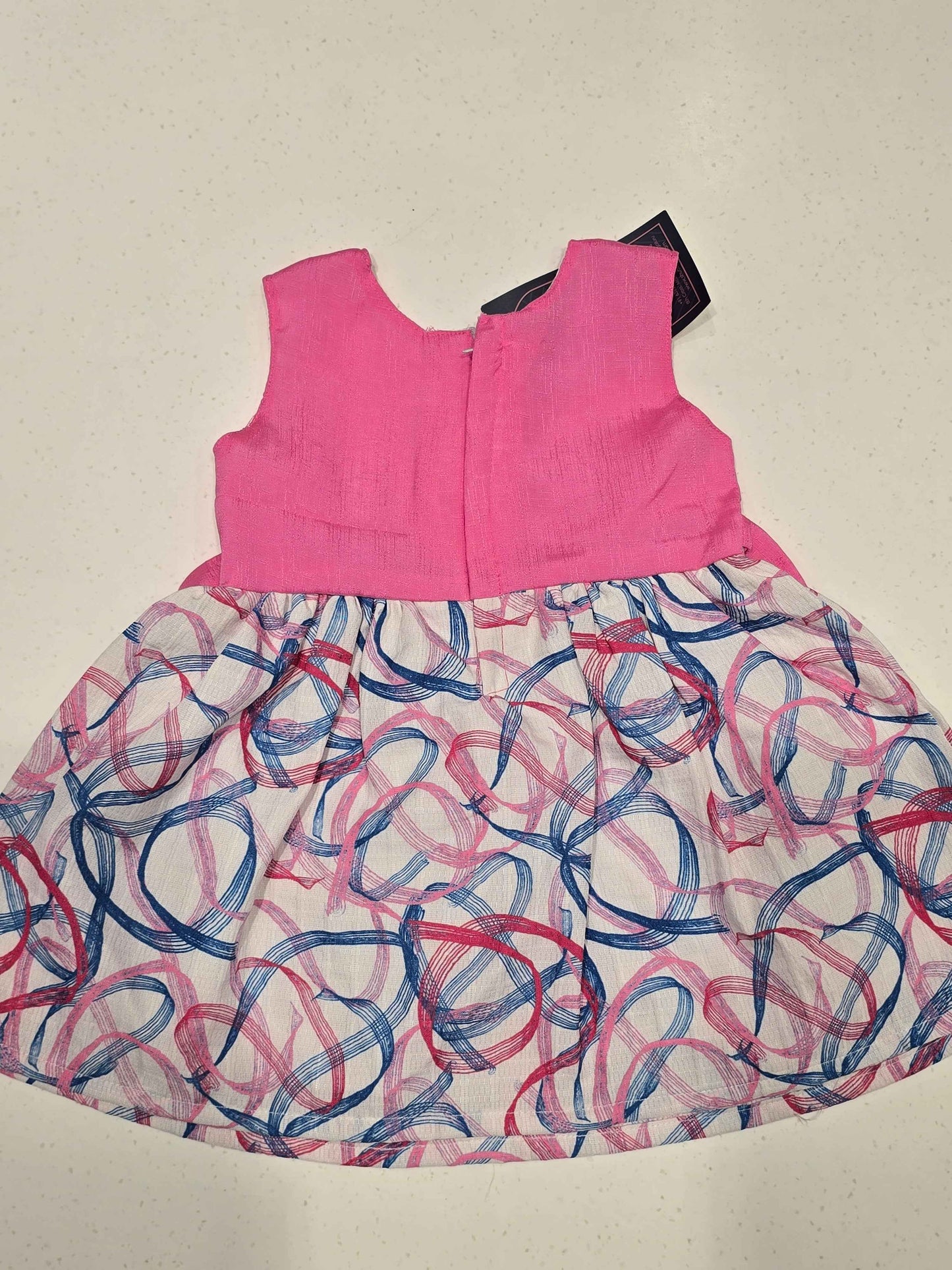 sleeveless cotton baby frock features a bright pink bodice paired with a contrasting white skirt decorated with abstract swirls. A light pink polka-dot bow accent highlights the satin-like ribbon waistband,