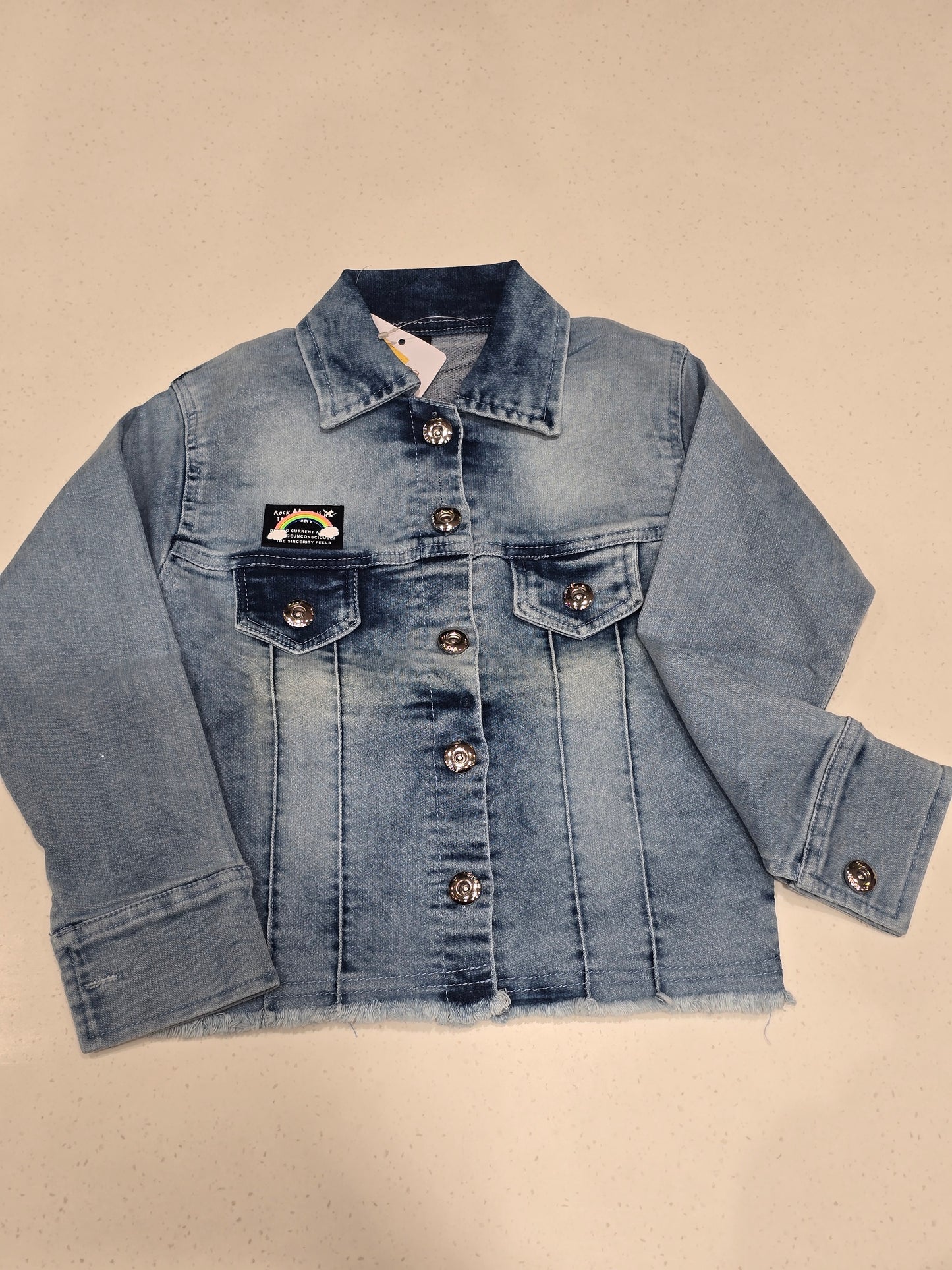 washed blue denim jacket featuring front flap pockets, a spread collar, custom decorative buttons, and a small embroidered chest patch.