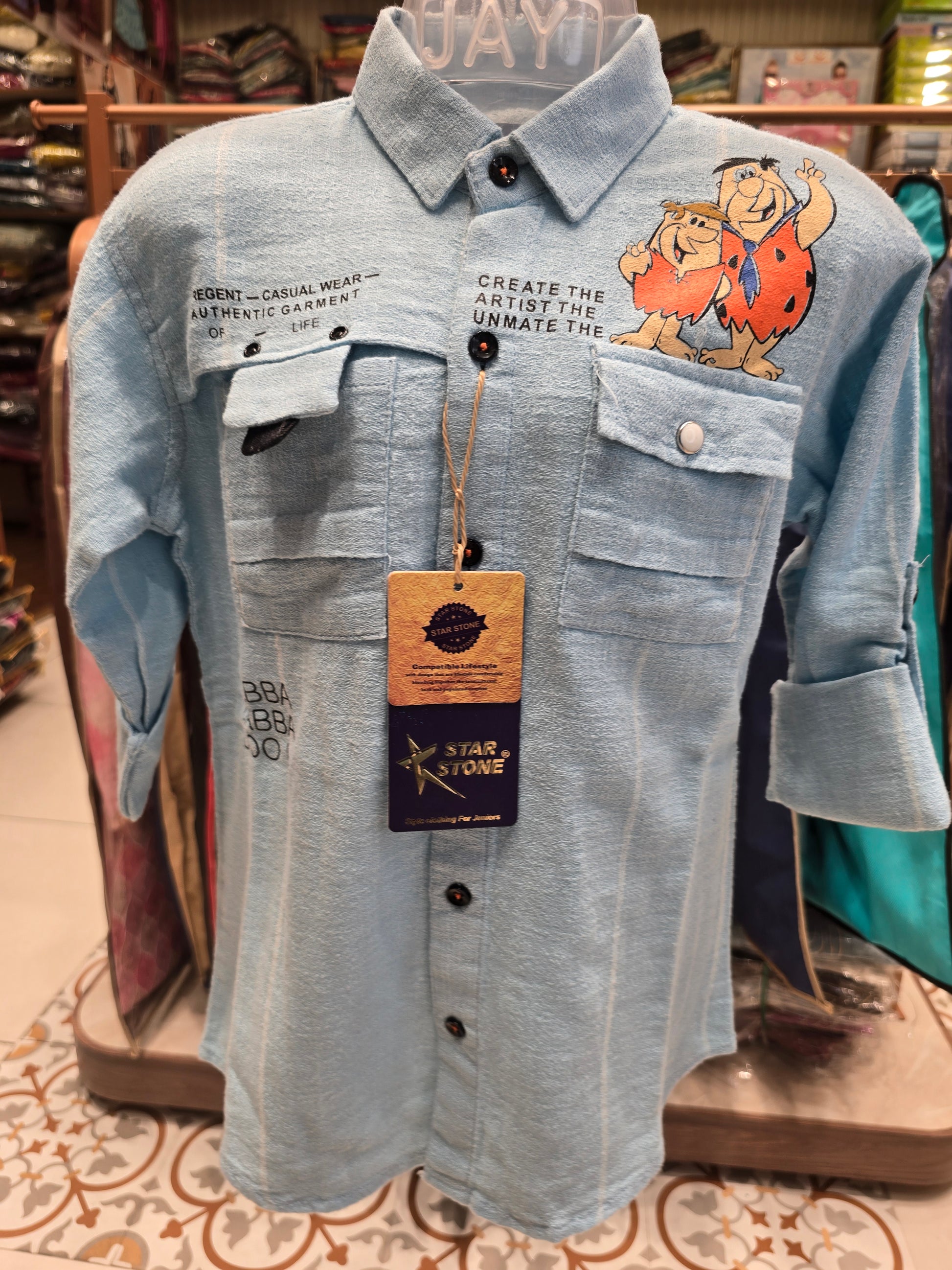 Light blue denim shirt with cartoon character design on a mannequin against a patterned wall.