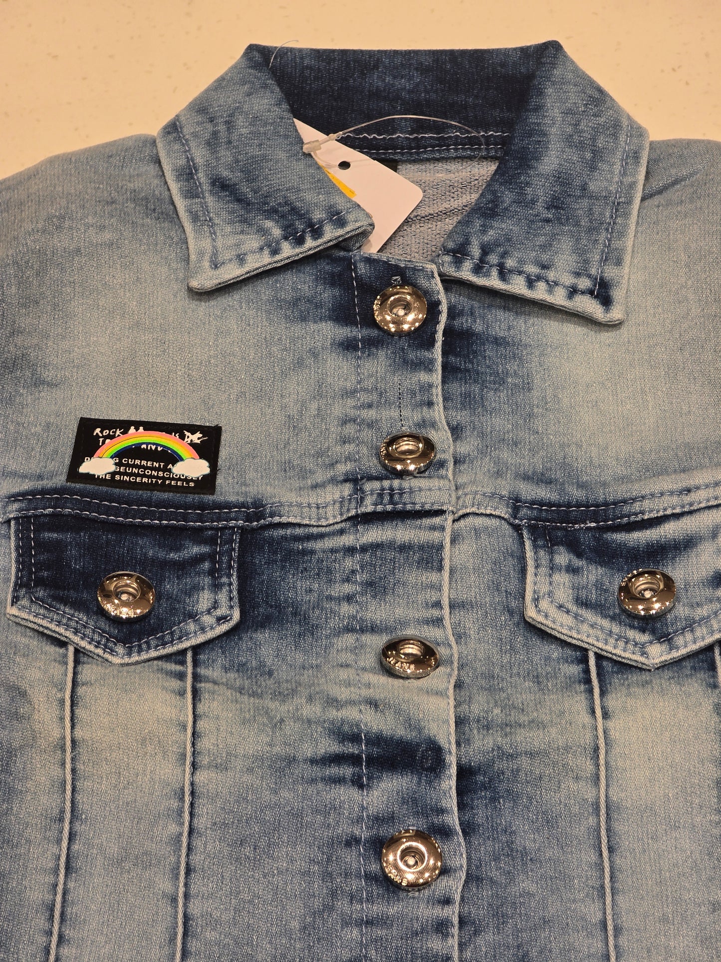 washed blue denim jacket featuring front flap pockets, a spread collar, custom decorative buttons, and a small embroidered chest patch.
